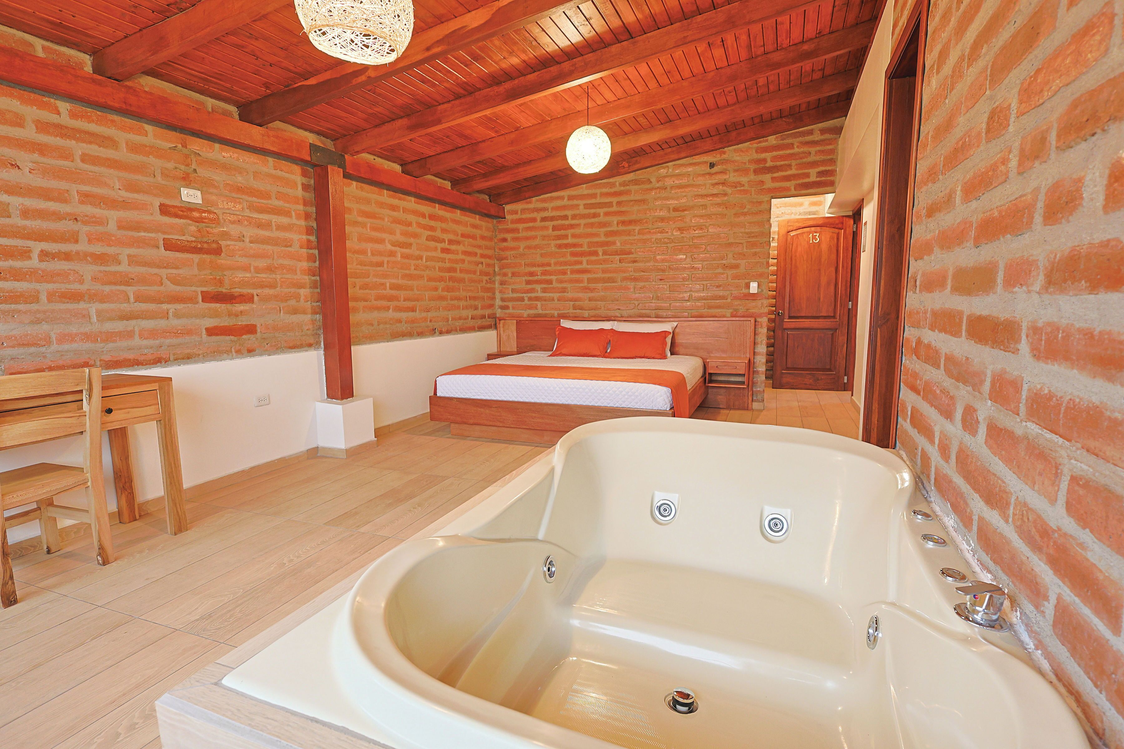 A room with a large bathtub, a bed with orange bedding, a wooden table, and a brick wall.