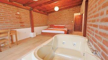 A room with a large bathtub, a bed with orange bedding, a wooden table, and a brick wall.