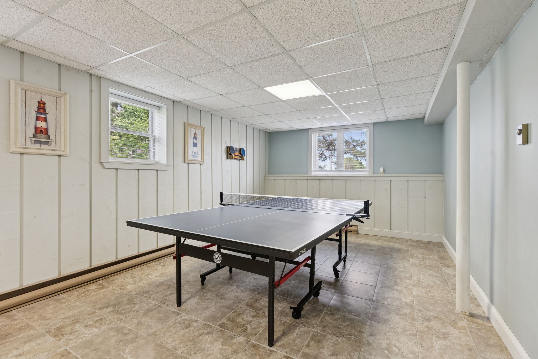 A ping pong table in a room with white paneled walls, two windows, and a ceiling with square tiles.