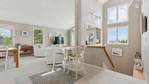 A spacious living area with a dining table, white chairs, a sofa, and a large window offering a view of the outdoors. - Drakes Island! Ocean Views!- Across from Drake's Island Beach! (Wells)