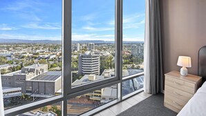 A hotel room with a large window offering a city view, a bed, a bedside table with a lamp, and a wooden dresser. - Comfortable Standard Studio in Surfers Paradise (Surfers Paradise)