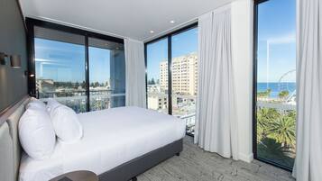 A modern hotel room with a large bed, a bedside table, and a view of the city and ocean through floor-to-ceiling windows.