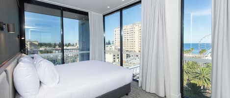 A modern hotel room with a large bed, a bedside table, and a view of the city and ocean through floor-to-ceiling windows.