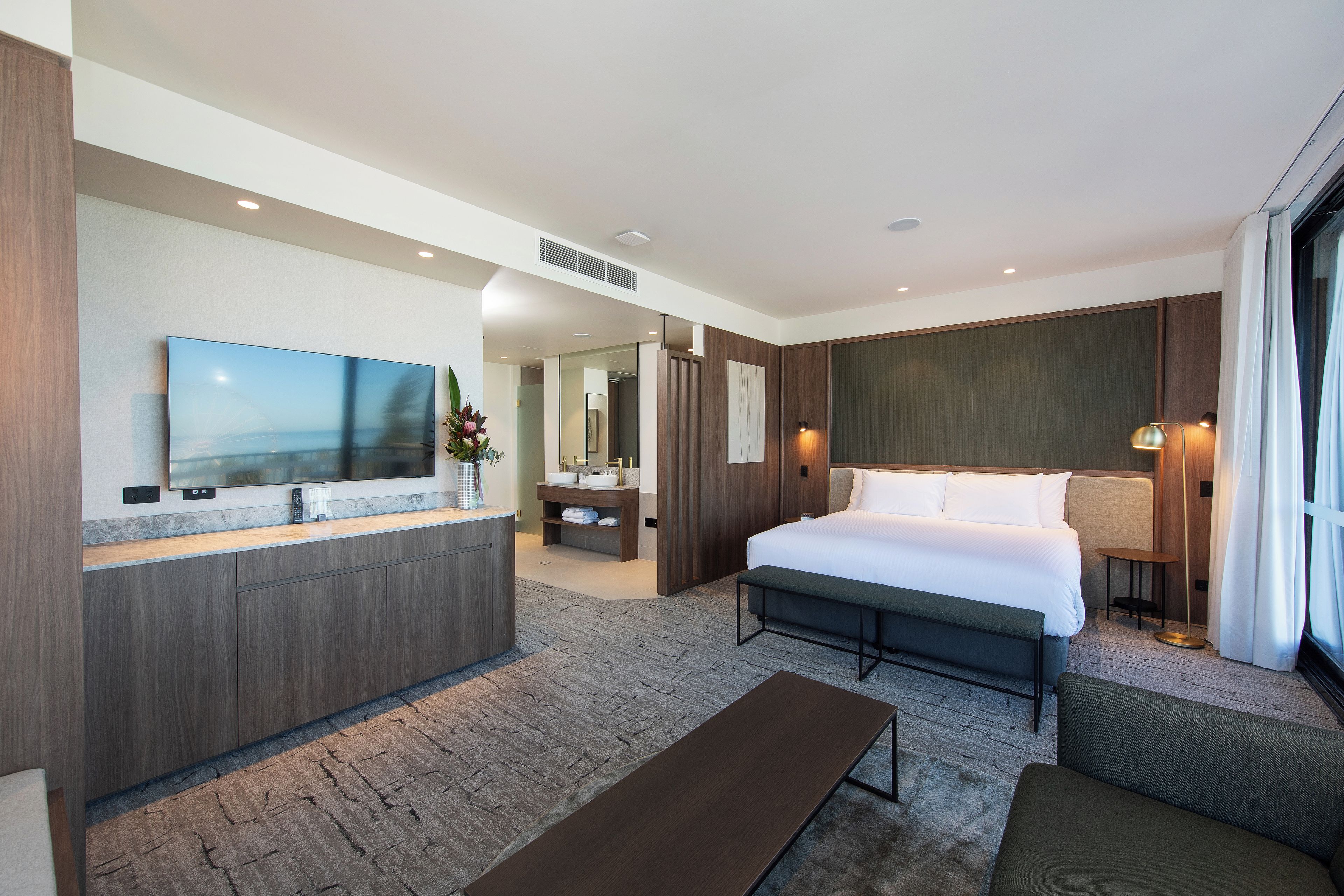 A modern hotel room with a large bed, a bedside table, a sofa, a TV, a bathroom with a mirror, and a large window.