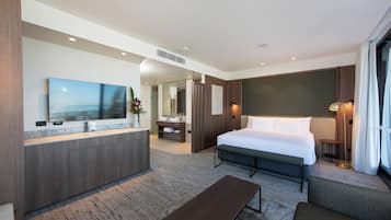 A modern hotel room with a large bed, a bedside table, a sofa, a TV, a bathroom with a mirror, and a large window.