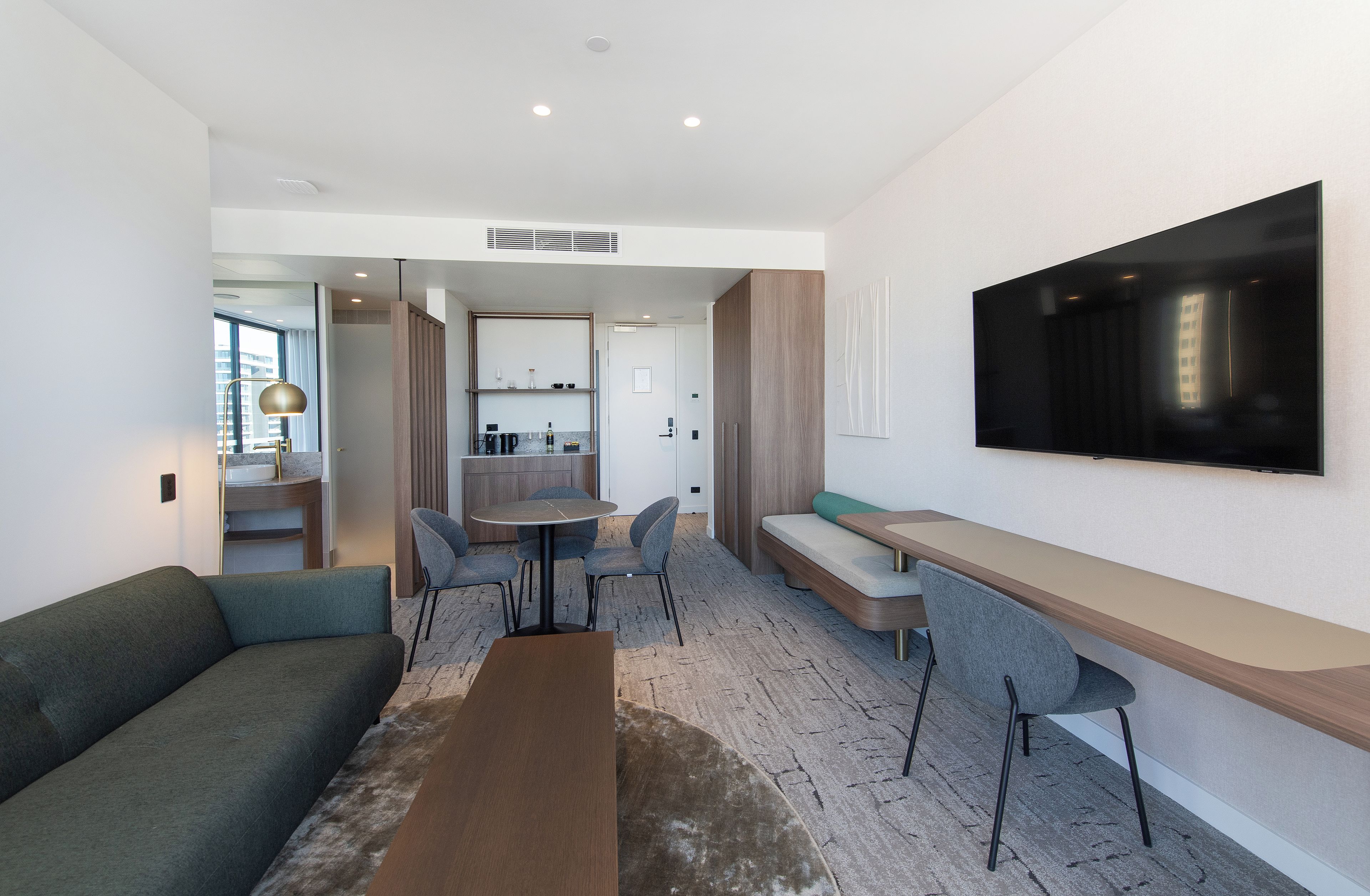 A modern hotel room with a sofa, a dining table, chairs, a bench, and a TV mounted on the wall.