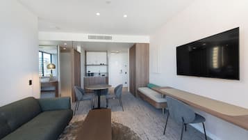 A modern hotel room with a sofa, a dining table, chairs, a bench, and a TV mounted on the wall.