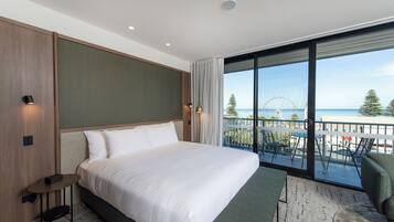 A modern hotel room with a large bed, a small table, and a balcony with a view of the sea and a Ferris wheel.