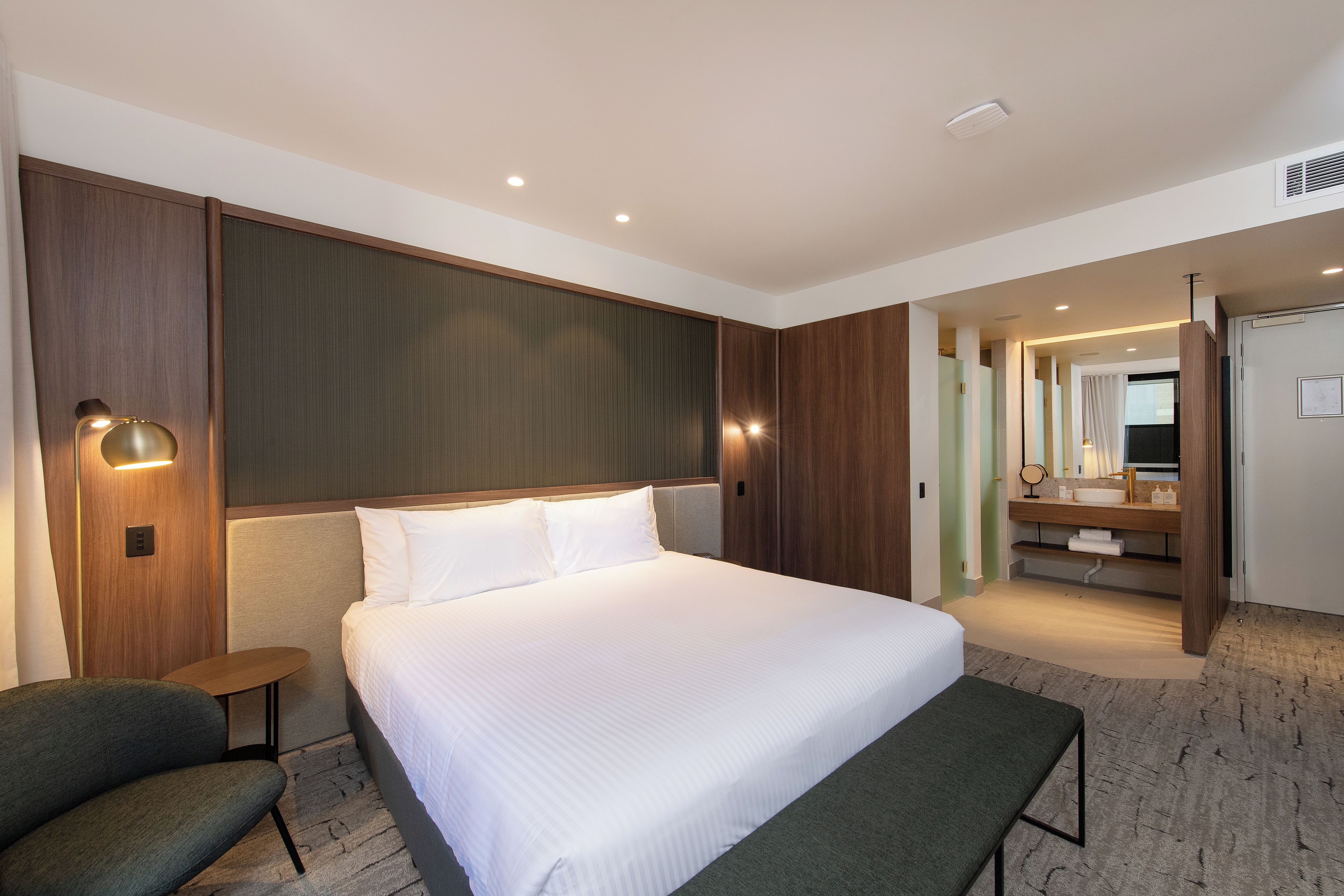 A hotel room with a large bed, a bedside table, a chair, a mirror, and a bathroom visible through an open door.