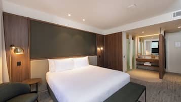 A hotel room with a large bed, a bedside table, a chair, a mirror, and a bathroom visible through an open door.