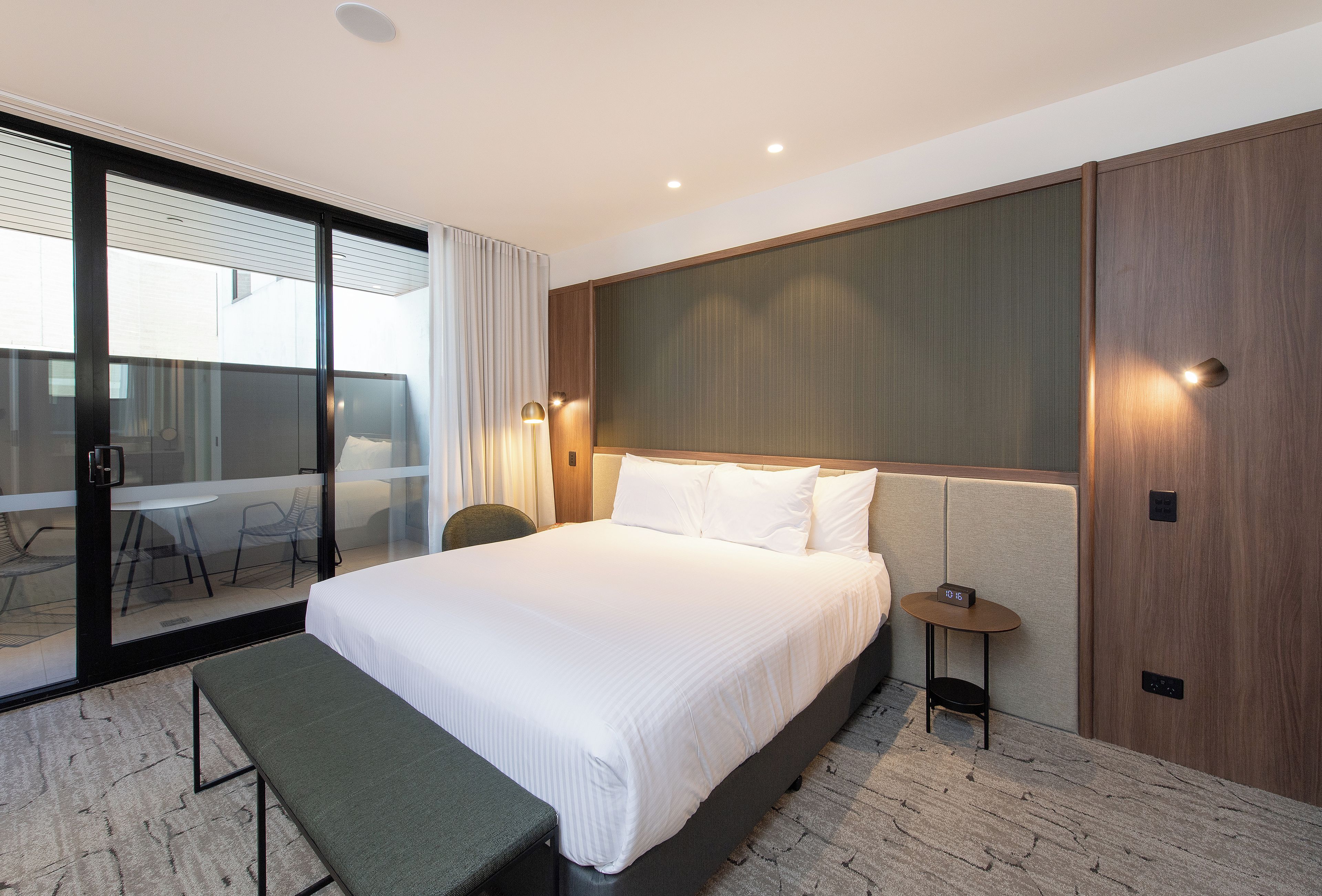 A hotel room with a large bed, a bench, a small table with a clock, and a sliding glass door leading to a balcony with a table and chairs.