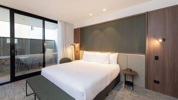 A hotel room with a large bed, a bench, a small table with a clock, and a sliding glass door leading to a balcony with a table and chairs.