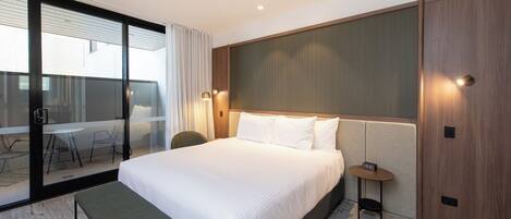 Habitación de hotel con una cama grande, un banco, una mesita con un reloj y un ventanal corredero que da a un balcón con mesa y sillas.