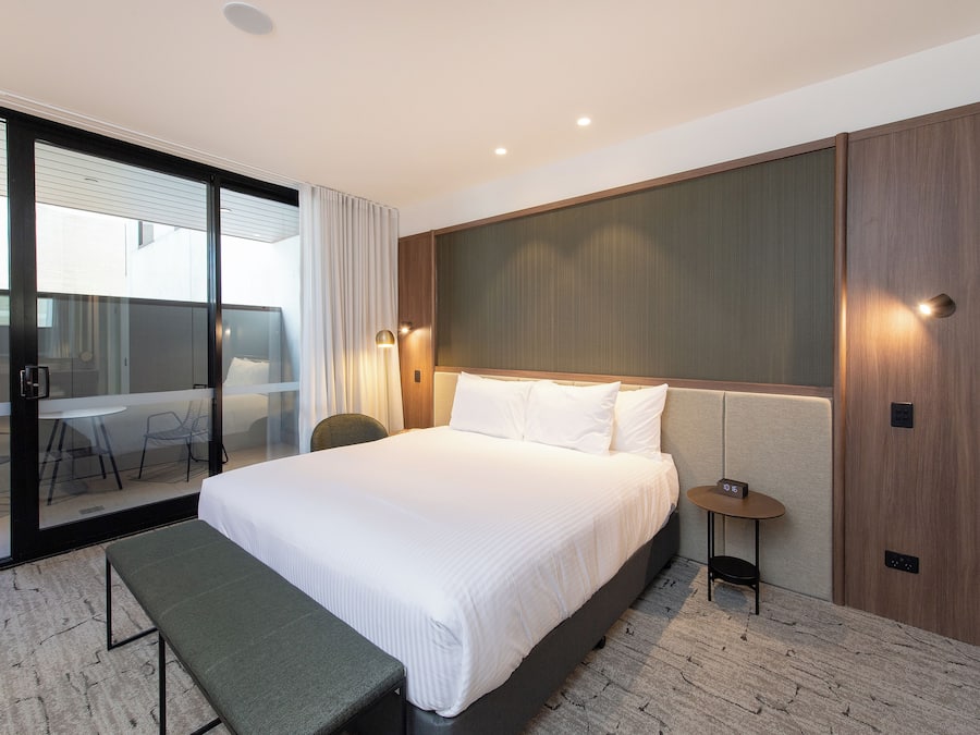 Quarto de hotel com uma cama grande, um banco, uma pequena mesa com relógio e uma porta de vidro deslizante para uma varanda com mesa e cadeiras.