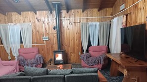 A cozy cabin interior with a wood-burning stove, two armchairs, a sofa, and a television on a wooden table. - Isla de Pino Huacho (Villarrica)