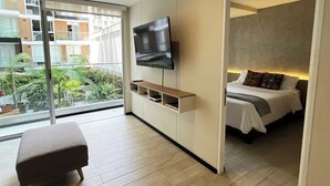 A modern hotel room with a flat-screen TV mounted on a wall, a comfortable sofa, a bed with a nightstand, and a balcony with a view of the city. - Coastal Blvd Apartment -Barranco (Lima)