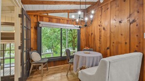A cozy room with wooden walls, a large window, a chandelier, and a table with chairs. - Riverbend Retreat (Berryville)