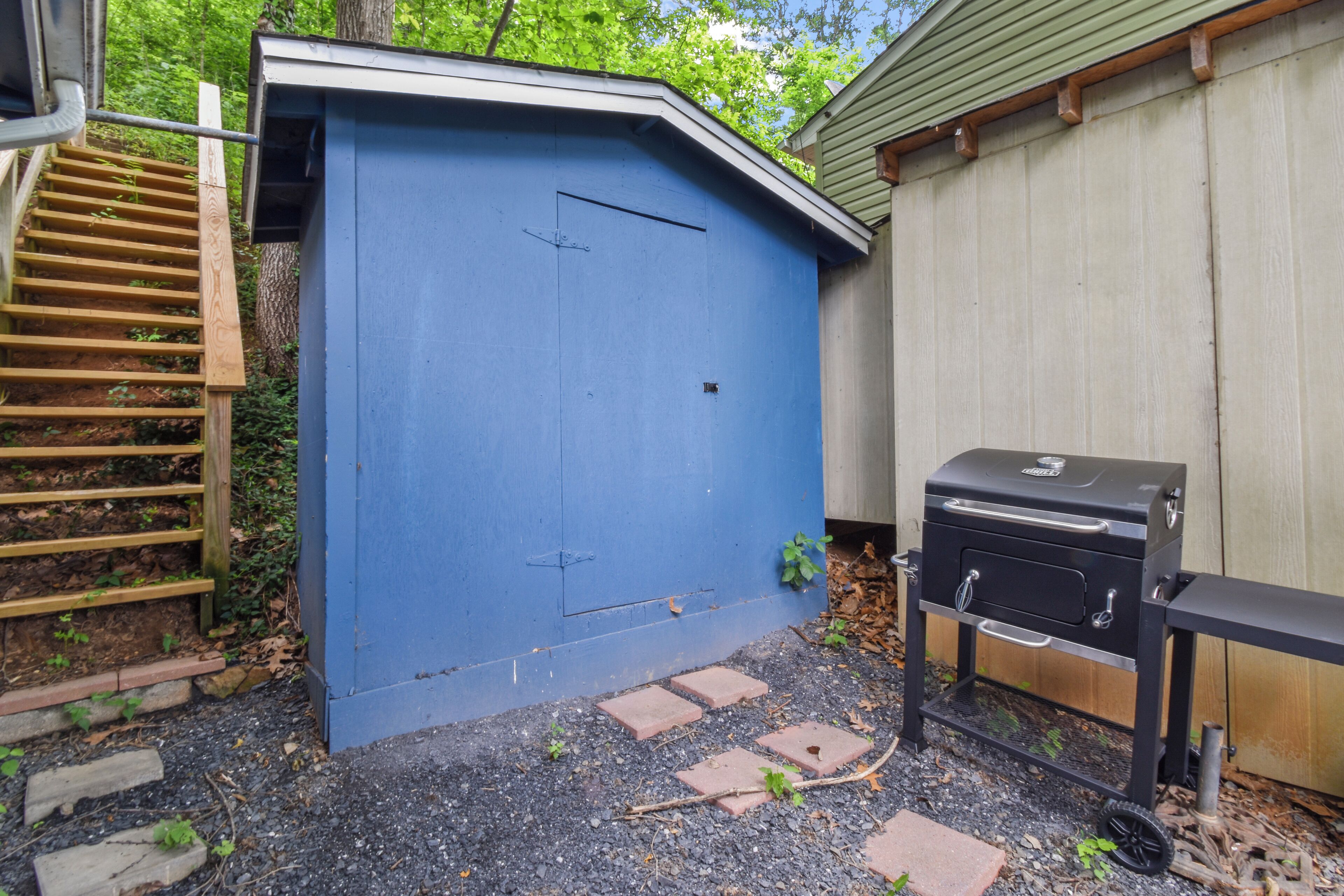 A blue shed with a wooden ladder, a barbecue grill, and a gravel path.