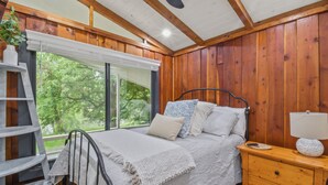 A bedroom with a bed, bedside table, lamp, and a window with a view of trees. - Riverbend Retreat (Berryville)