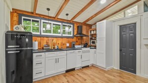 A kitchen with white cabinets, a black refrigerator, wooden flooring, and a window with a view of greenery. - Riverbend Retreat (Berryville)