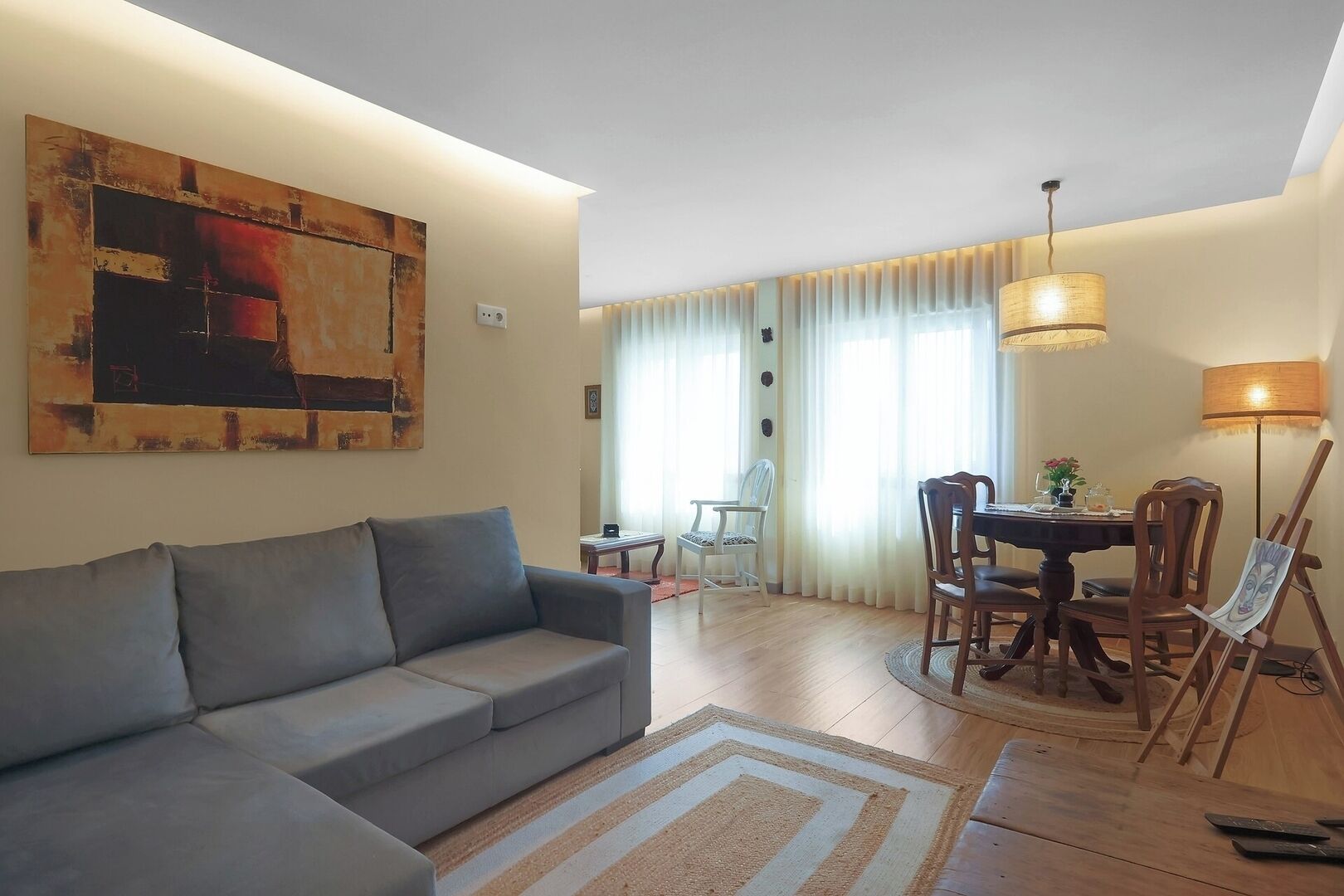 Ein Wohnzimmer mit grauem Sofa, einem Esstisch mit Stühlen, einem Bild an der Wand und einer Lampe.