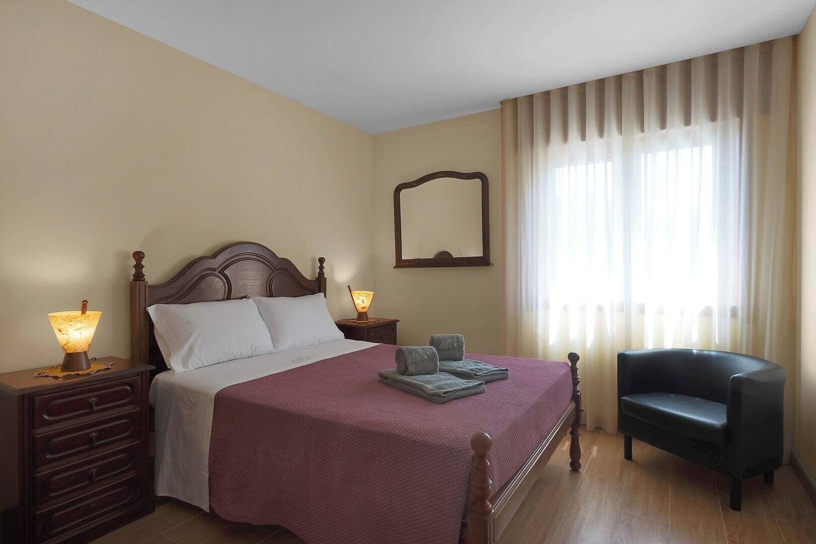 Une chambre à coucher bien rangée, avec un lit en bois, deux lampes de chevet, une chaise et une grande fenêtre avec des rideaux.