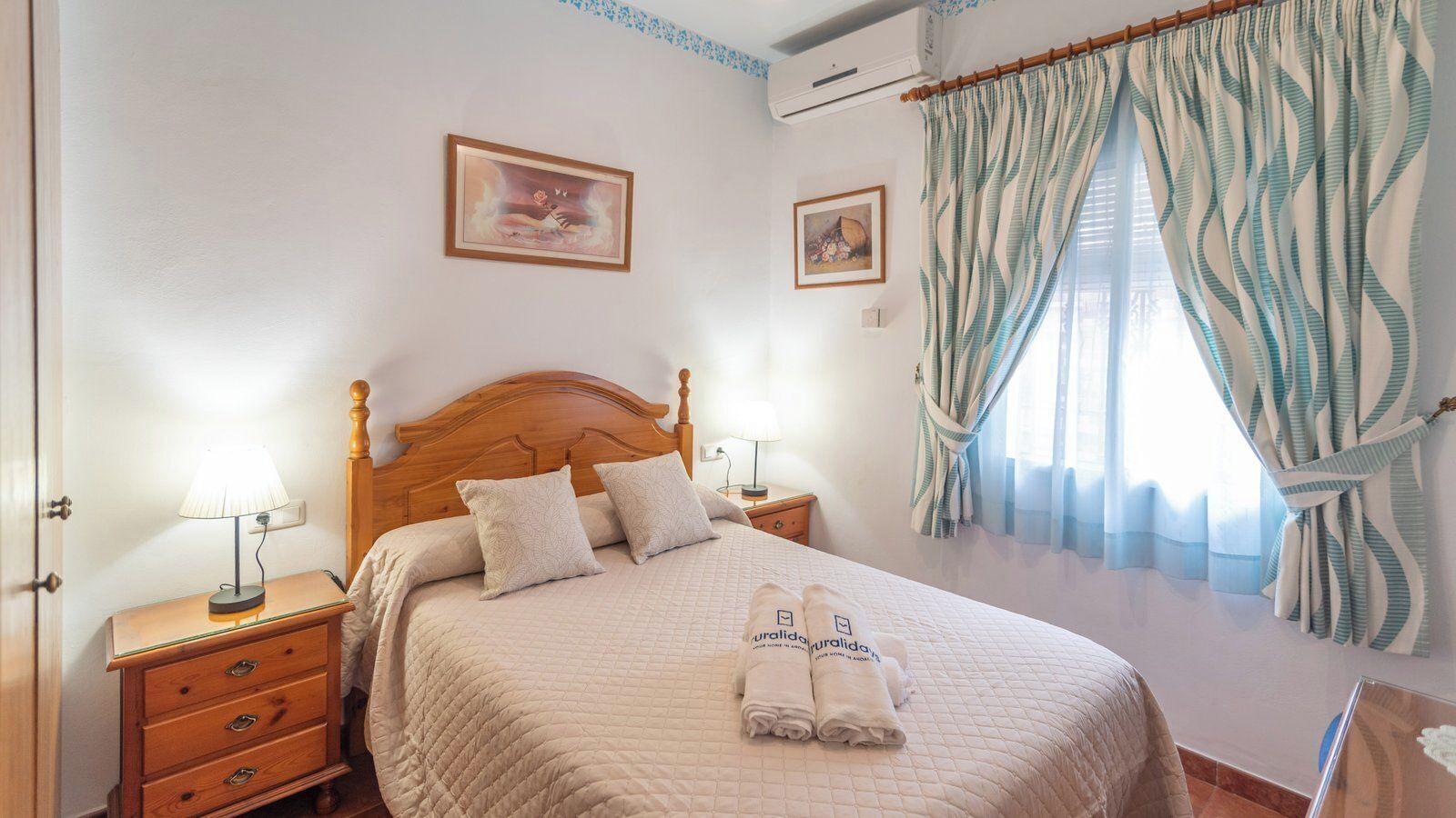 A neatly made bed with a quilted cover, two bedside tables with lamps, a wooden headboard, and a window with patterned curtains.