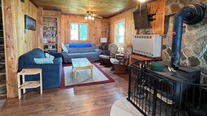 A cozy cabin interior with a stone wall, wooden ceiling, a fireplace, and a living area with a sofa and chairs.