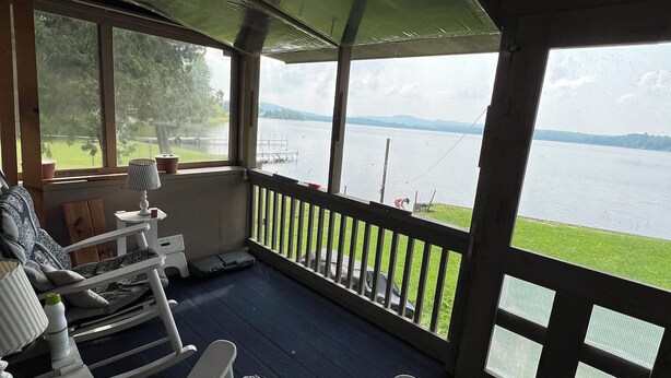 A covered porch with a view of a lake, a rocking chair, a table with a lamp, and a potted plant.