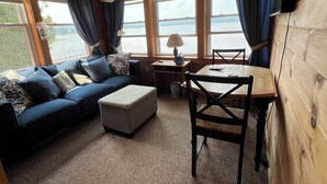 A cozy cabin interior with a wooden ceiling, a sofa set, a dining table, and large windows offering a view of the outdoors.
