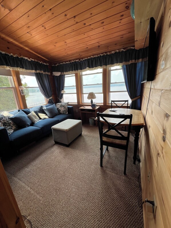 A cozy cabin interior with a wooden ceiling, a sofa set, a dining table, and large windows offering a view of the outdoors. - Charming 5-bedroom lake house on lake Salem in Derby, Vermont. (Derby)