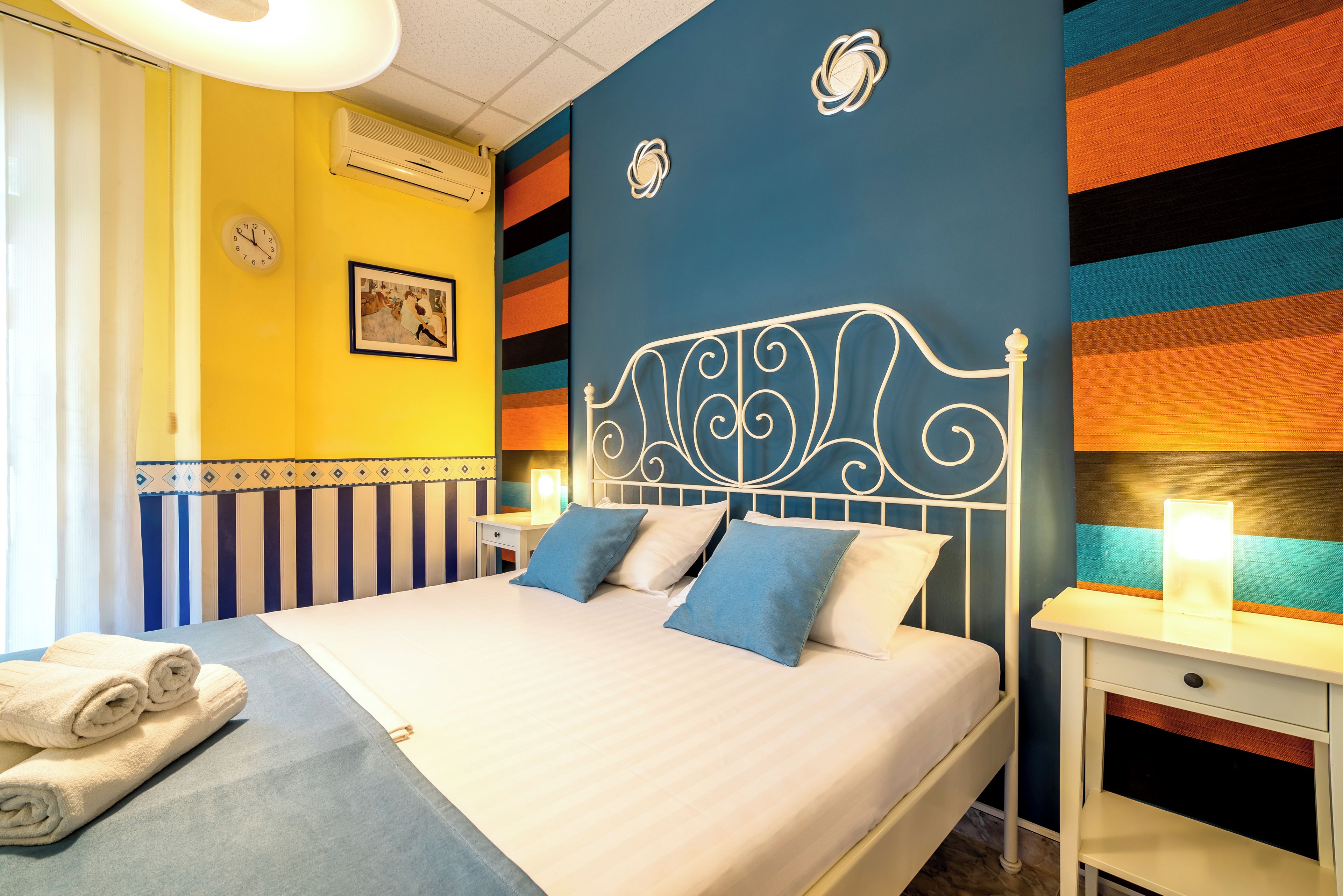 Un lit avec une tête de lit blanche, deux oreillers et une couvre-lit bleue. Il y a une table de chevet avec une lampe et un tableau encadré au mur.