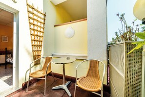 Een klein terras met rieten stoelen en een ronde tafel, een houten spijlen en een witte schelpdcoratie aan de muur.