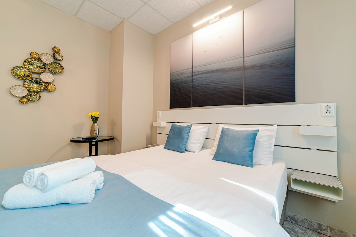 A neatly made bed with white linens and blue pillows, a wall art piece, and a small table with a vase of flowers.