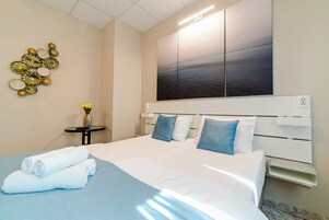 Una cama bien hecha con sábanas blancas y almohadas azules, una obra de arte en la pared y una mesita con un florero.
