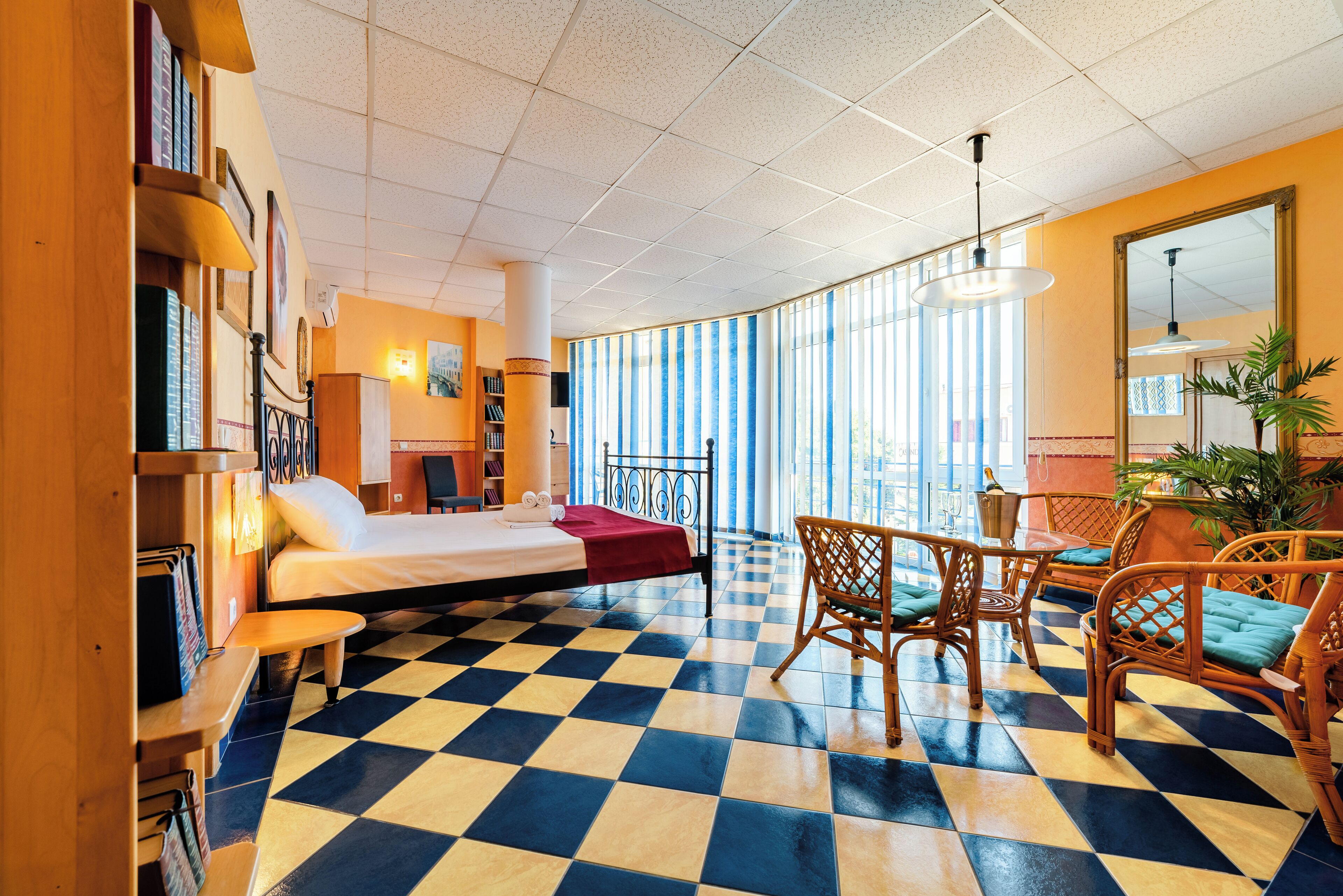 A hotel room with a checkered floor, a bed, a desk, and a dining area with wicker furniture.