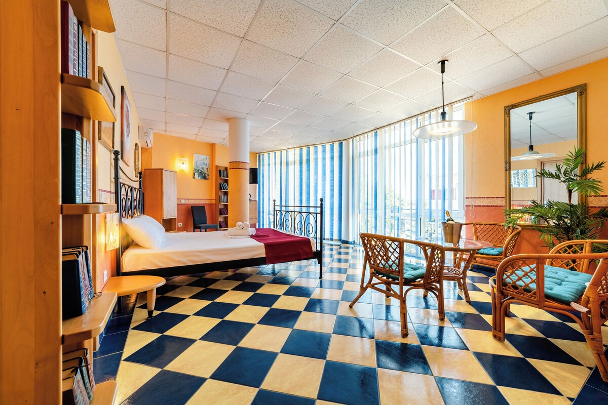 Een hotelkamer met een geruite vloer, een bed, een bureau, rieten stoelen en grote ramen.