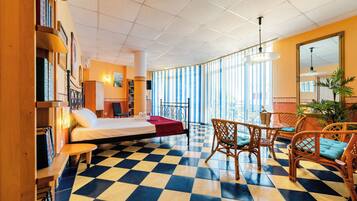 A hotel room with a checkered floor, a bed, a desk, wicker chairs, and large windows.