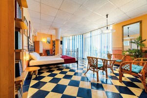 A hotel room with a checkered floor, a bed, a desk, wicker chairs, and large windows.