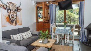 A living room with a grey sofa, a coffee table with a plant, a TV mounted on the wall, and a view of the outside through a door. - Buckleigh Lodge (Newton Abbot)