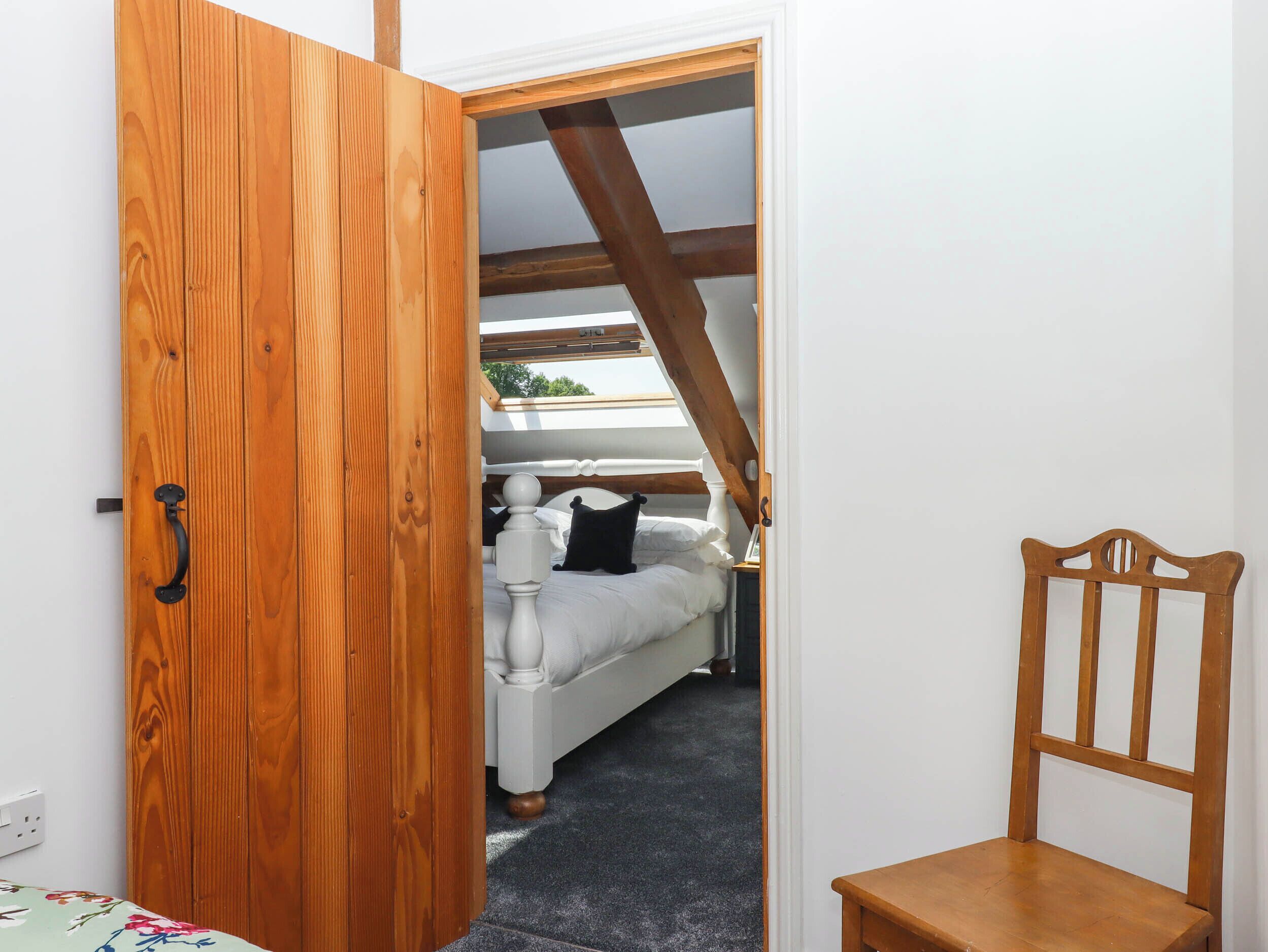 A bedroom with a wooden door, a bed, a wooden chair, and a sloped ceiling with wooden beams.