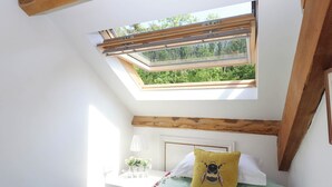 A bedroom with a skylight, a bed with a bee pillow, a bedside table with a lamp, and a window overlooking greenery. - Buckleigh Lodge (Newton Abbot)