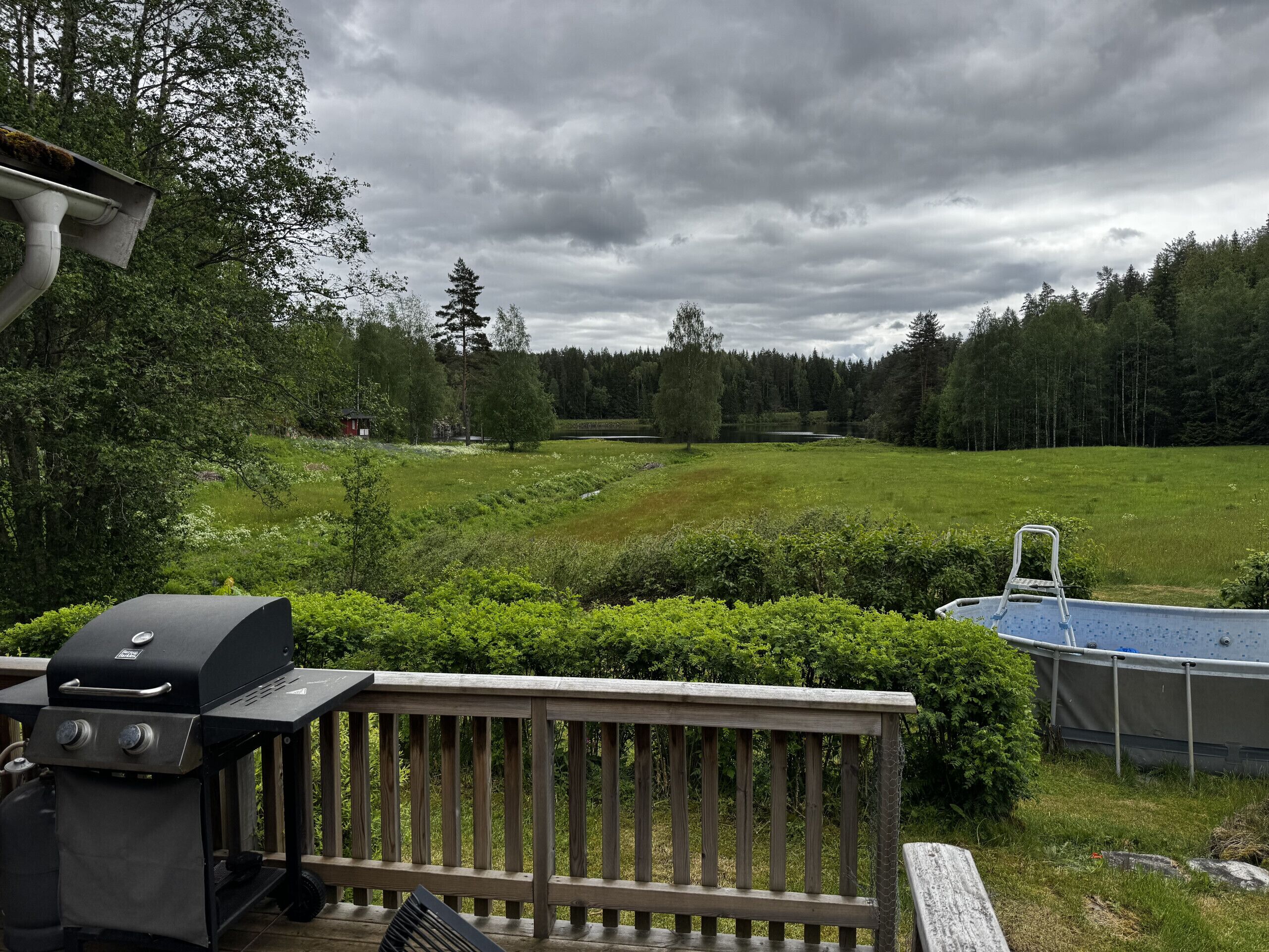 Une terrasse en bois avec un barbecue, une piscine et une vue sur un champ verdoyant et une forêt.