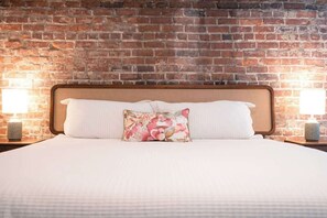 Un lit bien fait, agrémenté d’un coussin à motifs floraux, de deux lampes de chevet et d’un mur en briques en arrière-plan.