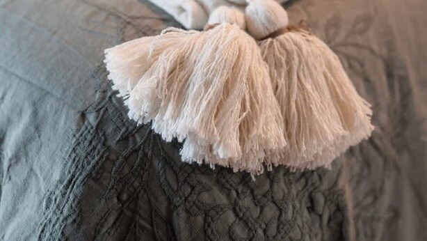 Uma cama bem arrumada com um edredom cinzento e um cobertor branco com borlas.