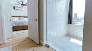 Habitación de hotel con cama, ventilador de techo, ventana con vistas a un paisaje urbano y pared de azulejos blancos.