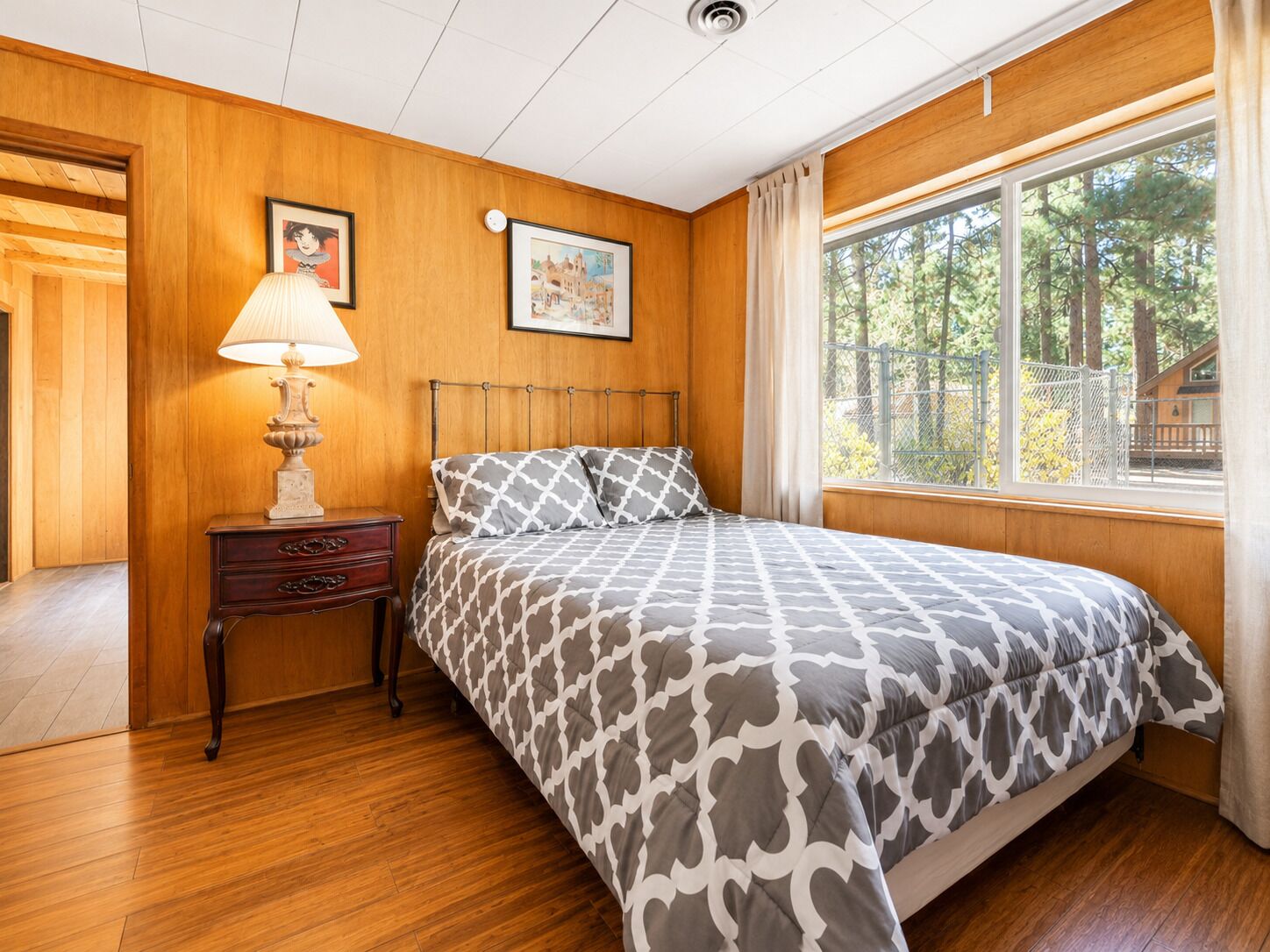 A bedroom with a wooden paneled wall, a bed with patterned bedding, a nightstand with a lamp, and a window with a view of trees.