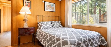 A bedroom with a wooden paneled wall, a bed with patterned bedding, a nightstand with a lamp, and a window with a view of trees.