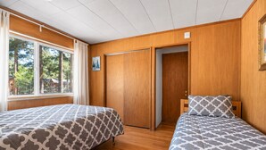 A bedroom with two beds, wooden flooring, a large window with curtains, and a door leading to another room.