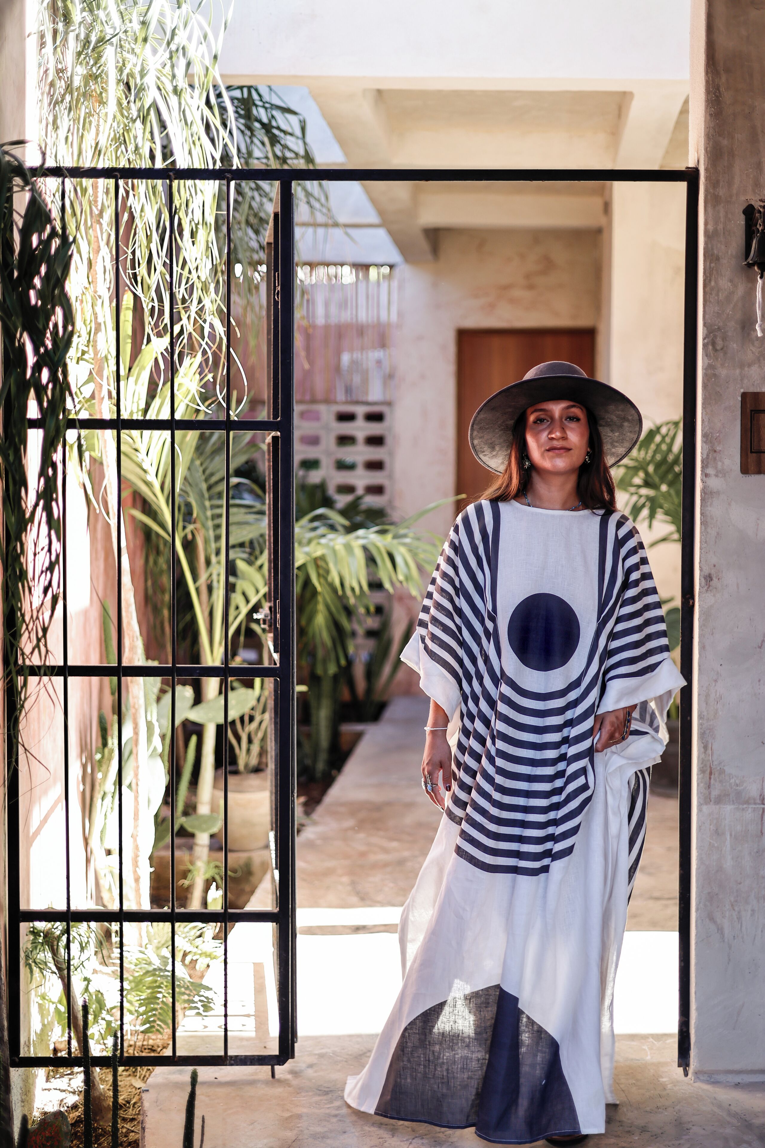 A person standing in a doorway with a striped garment and a hat, surrounded by greenery and a building exterior.